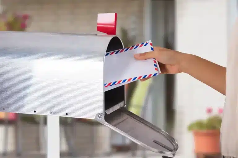 A person's hand is placing an airmail envelope, akin to a postcard template, into an open silver mailbox with a red flag. The background reveals a blurred outdoor setting.